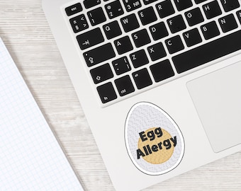 May include: An egg-shaped sticker with the words "Egg Allergy" in black text on a yellow circle, surrounded by a white and grey eggshell design. The sticker is on a laptop.