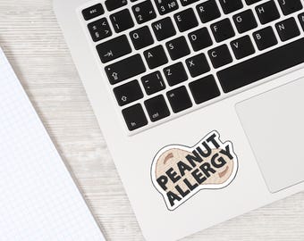 May include: A white laptop with a black keyboard and a sticker that says "PEANUT ALLERGY" in black text. The sticker is shaped like a peanut and has a tan background. A notebook is in the corner.