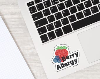 May include: A white sticker on a laptop with a red strawberry and blue blueberries, with the words "Berry Allergy" in black text. The sticker is on a white laptop, next to a notebook.