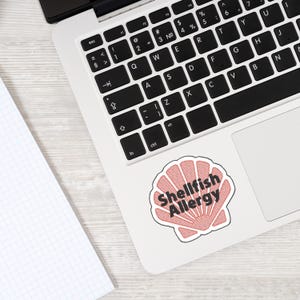 Può includere: Un adesivo con un design a conchiglia rossa e le parole "Shellfish Allergy" in testo nero. L'adesivo è su un laptop bianco, con una tastiera visibile.