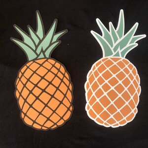Peut inclure: Deux découpes en forme d'ananas. Les ananas ont des corps orange avec un motif quadrillé et des sommets feuillus verts et vert clair. Une découpe a un contour noir, et l'autre a un contour blanc. Le fond est noir.