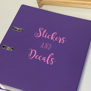 May include: A purple binder with silver metal rings. The cover features the pink text "Stickers AND Decals" in a cursive font. The binder is on a white surface.