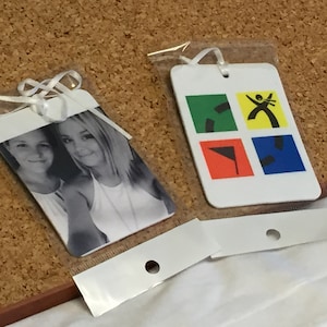 May include: Two white rectangular tags in clear plastic packaging. One tag features a black and white photo of two people. The other tag has a colorful graphic design with green, yellow, red, and blue squares. White ribbon is attached.
