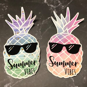 Peut inclure: Deux autocollants en forme d'ananas avec des lunettes de soleil et le texte "Summer Vibes". Les ananas ont des motifs aquarelles bleus, verts et violets, et roses, oranges et violets. Des paillettes dorées ajoutent des détails.