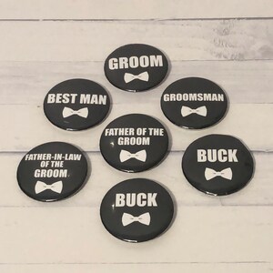 May include: A selection of black circular badges featuring white text and bow tie designs. The badges display phrases such as "Groom," "Best Man," "Groomsman," "Father of the Groom," and "Buck." These are likely for a wedding.