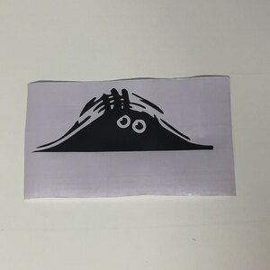 Puede incluir: Una pegatina de vinilo negro con un diseño de dibujos animados de una criatura asomándose por encima de una colina. La criatura tiene dos ojos blancos y un cuerpo negro. La pegatina está sobre un fondo blanco.