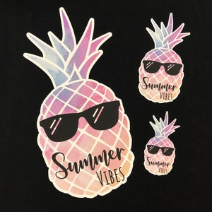 May include: Three pineapple stickers with sunglasses and the text "Summer Vibes". The pineapples have a watercolor design in shades of pink, purple, and blue. The stickers are on a black background.