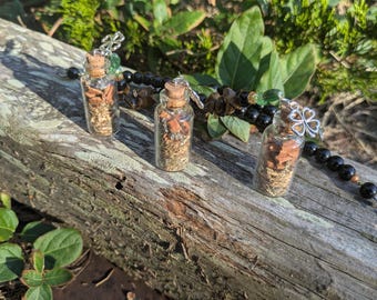 May include: Three small glass bottles with cork stoppers, filled with dried herbs and spices, are displayed on a weathered wooden surface. Each bottle has a silver charm attached. A black beaded necklace with green accents is also present, suggesting a natural or spiritual theme.