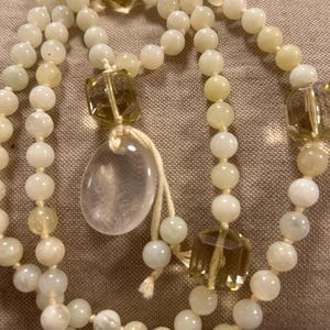 May include: A cream-coloured beaded necklace with a clear, teardrop-shaped pendant and several faceted, amber-coloured crystal accents. The necklace features round beads and is displayed on a neutral-toned surface.
