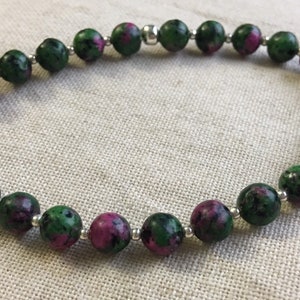 May include: A bracelet made of green and magenta gemstone beads, interspersed with small silver-coloured spacers. The beads are round and polished. The bracelet is presented on a neutral fabric background.
