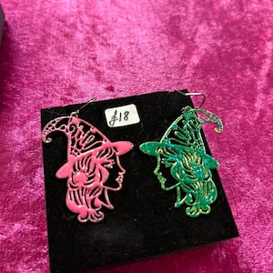 May include: A pair of witch-themed earrings, one pink and one green, with glitter accents. The earrings are displayed on a black background with a price tag that reads "£18".