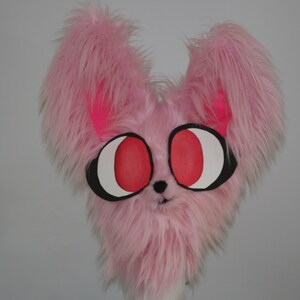 May include: Pink furry plush toy with large red eyes and black eye outlines. The toy has a small black nose and a pink mouth.