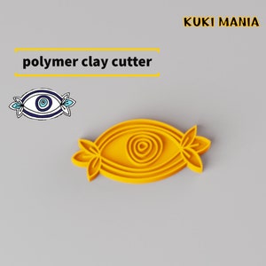 Könnte beinhalten: Gelber Kunststoff-Polymer Clay Cutter in Form eines bösen Auges mit einem Wirbelmuster in der Mitte. Der Cutter hat zwei kleine Blätter auf jeder Seite.