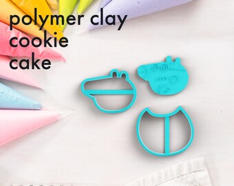 May include: Three blue plastic cookie cutters shaped like a pig, a crocodile, and a circle with a line down the middle. The cutters are on a white surface with a white and black striped towel in the bottom right corner. The text "polymer clay cookie cake" is written in black at the top of the image.