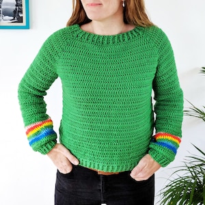 Könnte beinhalten: Ein grüner Häkelpullover mit Regenbogenstreifen an den Ärmeln. Der Pullover hat einen runden Ausschnitt und lange Ärmel.