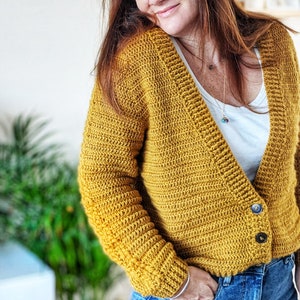 Può includere: Un cardigan in crochet giallo senape con scollo a V e chiusura con bottone. Il cardigan ha maniche lunghe e una texture a coste.
