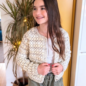 Puede incluir: Un cárdigan de crochet blanco con cierre de botón. El cárdigan tiene un diseño texturizado y abultado y se lleva sobre un top blanco sin mangas y pantalones verdes.