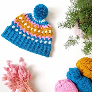 Può includere: Un berretto blu all'uncinetto con un pon pon bianco. Il berretto ha un motivo geometrico colorato in rosa, bianco e giallo.