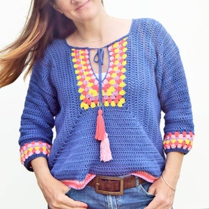 Puede incluir: Una blusa de crochet azul con un patrón geométrico colorido alrededor del escote y las mangas. La blusa tiene un lazo en el escote y una borla.