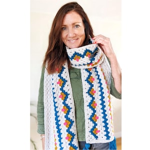 May include: A white crocheted scarf with blue, yellow, and pink geometric patterns. The scarf is worn by a person with long brown hair.