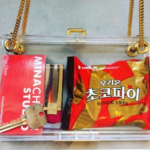 May include: A clear plastic clutch bag with a gold chain strap. Inside the bag are a red and white business card, a gold and red lipstick, and a red and gold bag of chips with the text "오리온 초코파이 SINCE 1974".