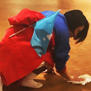 May include: A person wearing a red and blue costume with a white flag is kneeling on a wooden floor. The person is cleaning the floor with a white cloth.
