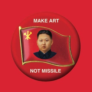May include: A red button with the text "MAKE ART" above and "NOT MISSILE" below a graphic of a flag with a portrait of a man. The button has a gold border and is set against a solid red background.