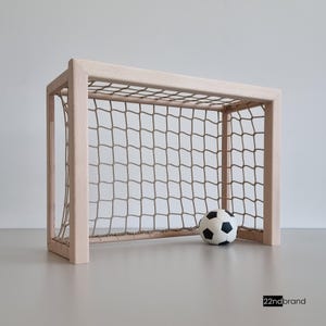 Könnte beinhalten: Ein Miniatur-Fußballtor aus hellem Holz mit beigem Netz. Ein schwarz-weißer Fußball liegt vor dem Tor. Das Tor ist ein Spielzeug oder Dekorationsartikel.