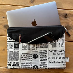 Könnte beinhalten: Eine schwarze Laptoptasche mit Reißverschluss. Die Tasche ist aus schwarzem Stoff mit einem Zeitungsdruckdesign. Der Zeitungsdruck zeigt Schlagzeilen wie "Overseas Crisis Stateside" und "Skirmish Between Two". Die Tasche ist mit einem Laptop und einem Tablet darin abgebildet.