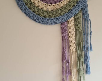 Puede incluir: Un tapiz de pared de macramé hecho a mano con un diseño de semicírculo. La pieza presenta una barra de madera y una paleta de colores azul, verde, morado y crema. El tapiz tiene flecos anudados y borlas.