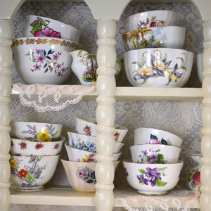 May include: A white wooden cabinet with multiple shelves filled with vintage teacups and saucers. The teacups are decorated with floral patterns in various colors, including pink, purple, yellow, and blue.