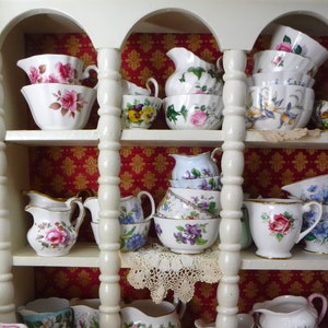 May include: A white wooden cabinet with multiple shelves filled with a collection of vintage teacups and saucers. The teacups are decorated with floral patterns in shades of pink, blue, and yellow. The cabinet has a red and gold patterned wallpaper background.