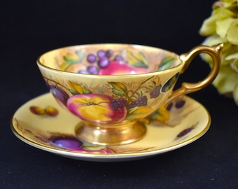 Può includere: Una tazza da tè e un piattino gialli con un disegno dipinto a mano di frutta, tra cui prugne, uva e mele. La tazza e il piattino hanno un bordo dorato.