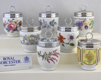 May include: Set of eight porcelain egg coddlers with floral designs and silver lids. The coddlers are arranged on a white tiered stand. The stand has a small sign that reads "Royal Worcester. Made in England."