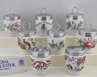 May include: Set of eight Royal Worcester egg coddlers with silver lids and various floral and bird designs. The coddlers are displayed on a white tiered stand. The Royal Worcester logo and "Made in England" are visible on a small white rectangular sign.