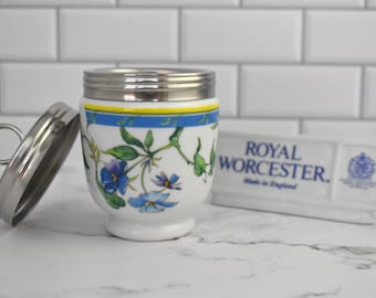 May include: White porcelain egg cup with a blue and yellow floral design. The egg cup has a stainless steel lid with a handle. The lid is open and the egg cup is sitting on a white countertop. The text "ROYAL WORCESTER. Made in England." is visible on a white rectangular plate.