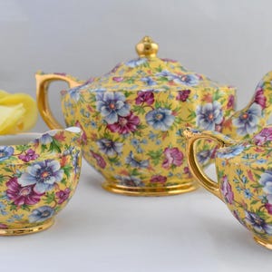May include: A yellow tea set with a floral pattern. The set includes a teapot, creamer, and sugar bowl. The floral pattern features pink, purple, and blue flowers on a yellow background.