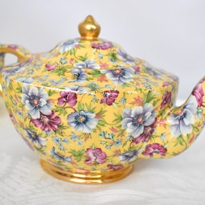 May include: A yellow teapot with a floral pattern. The teapot has a gold trim and a lid with a gold knob. The flowers are in shades of pink, blue, and white.