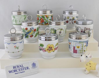 May include: A collection of white porcelain egg cups with silver lids. Each cup features a unique floral or fruit design, including apples, berries, and butterflies. The Royal Worcester logo is visible in the foreground. Made in England.