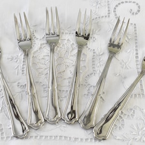 May include: Six silver forks with ornate handles are arranged in a fan shape on a white lace tablecloth.