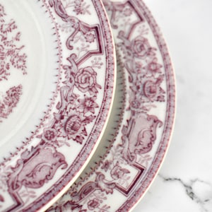 May include: Two white ceramic plates with a burgundy floral and architectural design. The plates have a detailed border with flowers and decorative elements. The plates are on a white marble surface.