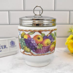 May include: A white porcelain egg coddler with a silver-tone metal lid featuring a ring handle. The body is decorated with a colorful fruit pattern including grapes, apples, and plums. A white rectangular plaque reads ROYAL WORCESTER. Made in England.