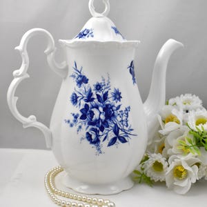 May include: A white porcelain teapot featuring a blue floral pattern on the body and lid. The teapot has a curved handle, a spout, and a scalloped base. The lid is topped with a decorative loop handle. The design includes detailed blue roses.