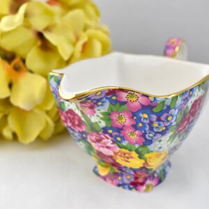 May include: A floral patterned creamer with a gold rim and handle. The creamer features a vibrant design of pink, purple, and yellow flowers on a blue background. A bouquet of yellow flowers is in the background.