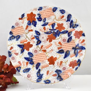 May include: A white porcelain plate with a scalloped edge features a floral pattern in shades of red, blue, and gold. The design includes stylized flowers, leaves, and geometric shapes.