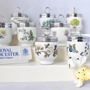 May include: A set of eight Royal Worcester egg coddlers with various floral and bird designs. The coddlers are white porcelain with silver lids. The coddlers are arranged on a white tiered stand. The stand has a Royal Worcester label with the text "Made in England".