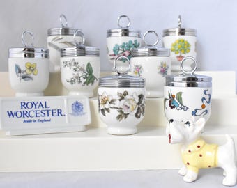 May include: A set of eight Royal Worcester egg coddlers with various floral and bird designs. The coddlers are white porcelain with silver lids. The coddlers are arranged on a white tiered stand. The stand has a Royal Worcester label with the text "Made in England".