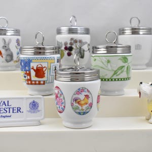 May include: A collection of Royal Worcester egg cups with silver lids, each decorated with unique designs including a rabbit, flowers, and a rooster. A small white dog figurine in a yellow jacket is also present. The Royal Worcester logo is visible.