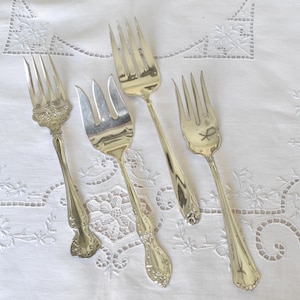 May include: Four silver forks with ornate handles. The forks are arranged on a white lace tablecloth.