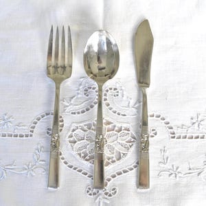 May include: A set of vintage silverware, including a fork, spoon, and butter knife, arranged on a white, embroidered tablecloth. The silverware is silver-toned with decorative floral detailing on the handles. The tablecloth has a delicate, lacy pattern.